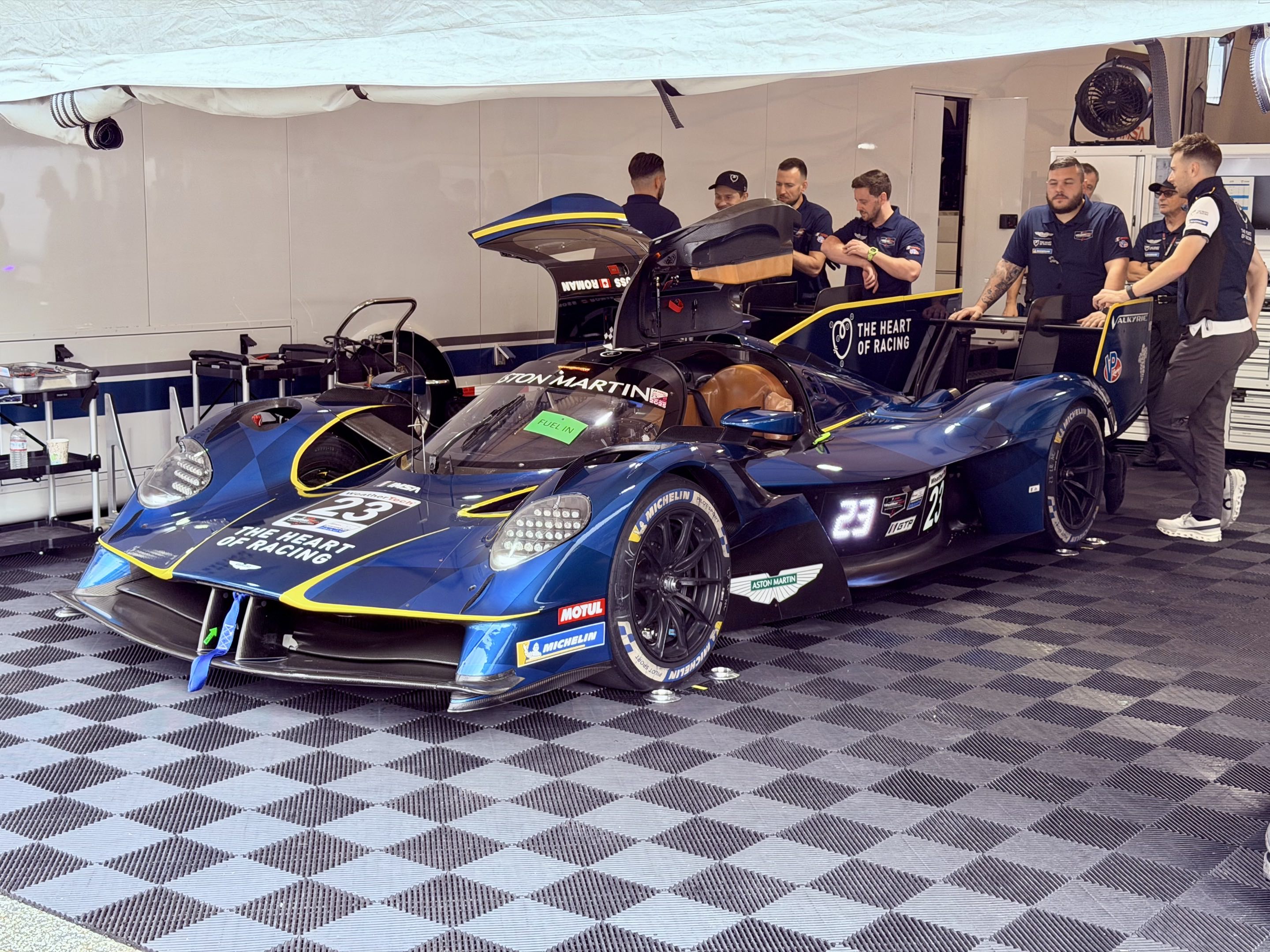 An Aston Martin Valkyrie AMR-LMH in its garage with its doors up and the team standing around behind it